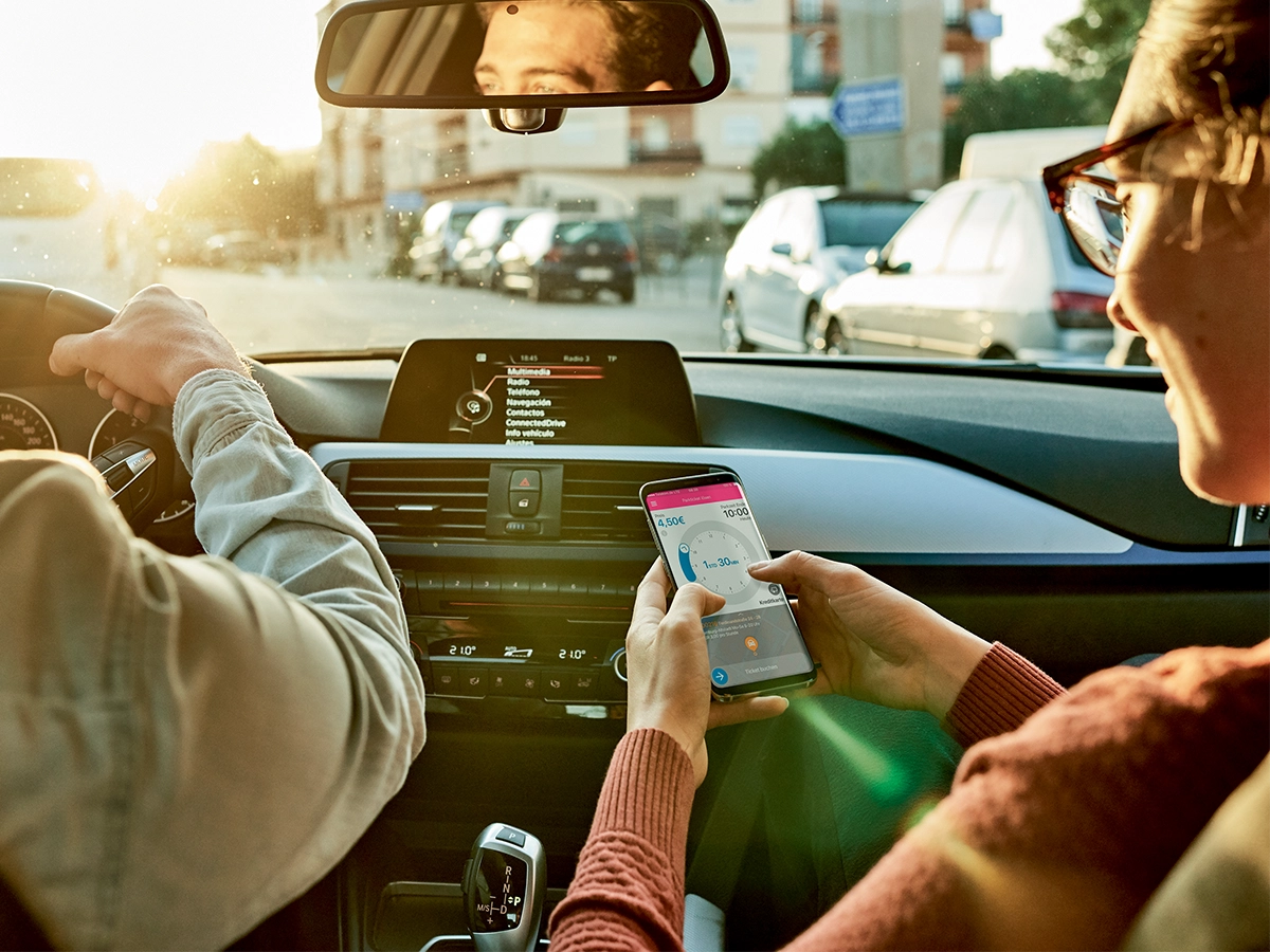 Zwei Personen sitzen in einem Auto. Die rechte Person hält ein Smartphone in den Händen.