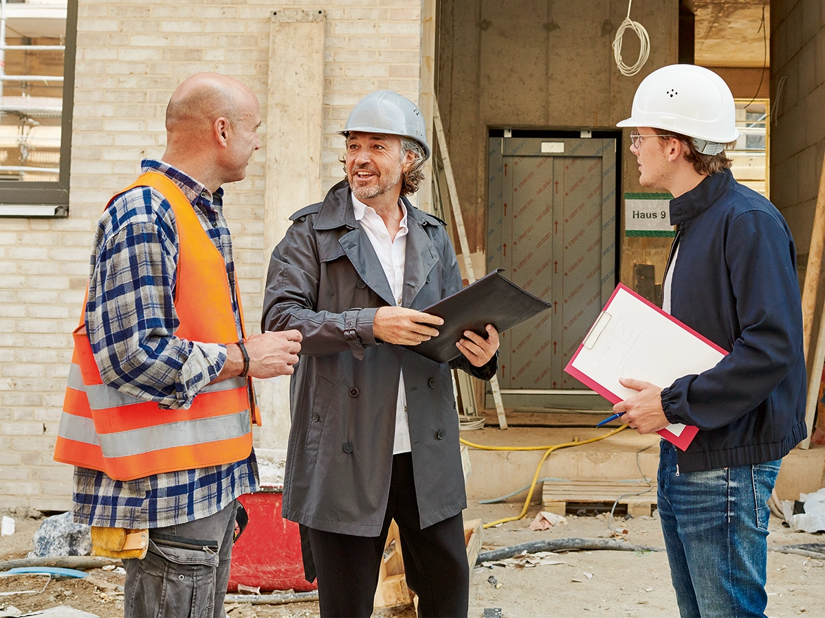 Drei Männer besprechen etwas auf einer Baustelle. Zwei davon tragen Klemmbretter mit sich und haben Helme auf dem Kopf.