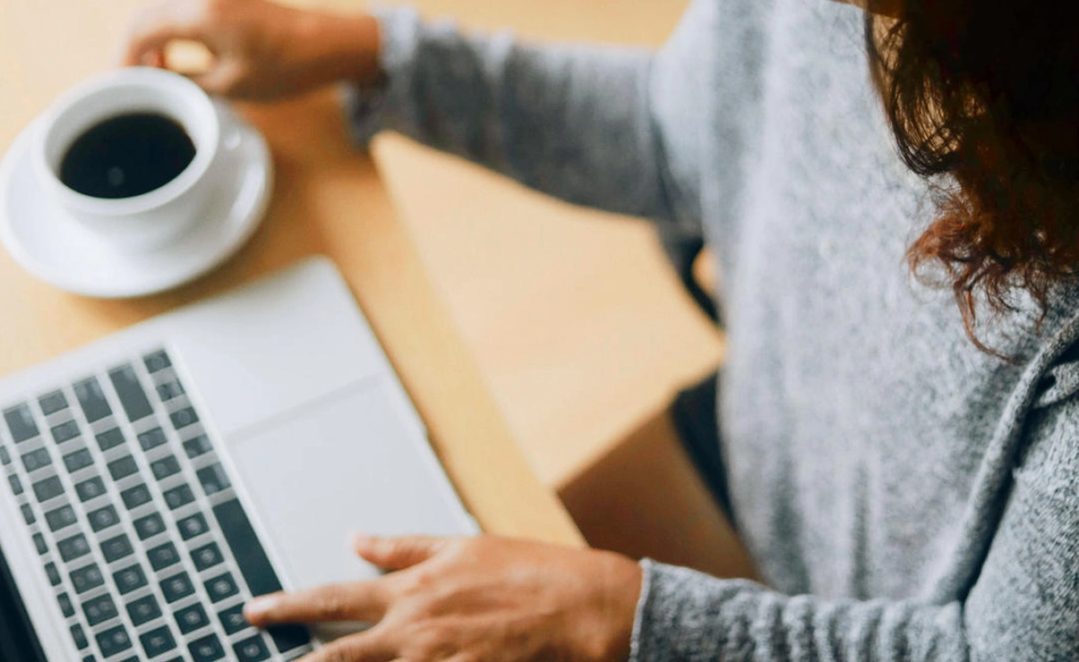 Das Bild zeigt eine Person, die an einem Laptop sitzt und daneben steht eine Tasse Kaffee. Das Bild ist aus der Vogelperspektive.