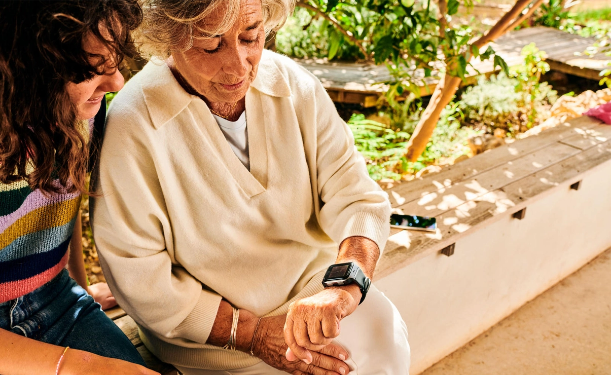 Eine ältere Frau und eine jüngere Frau schauen auf die Smartwatch der älteren Frau. Sie sitzen im Freien auf einer Bank.