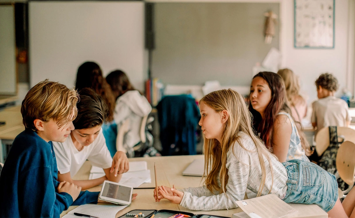 Das Bild zeigt eine Schulklasse, wo mehrere Kinder an Tischen sitzen. Im Fokus stehen vier Kinder.