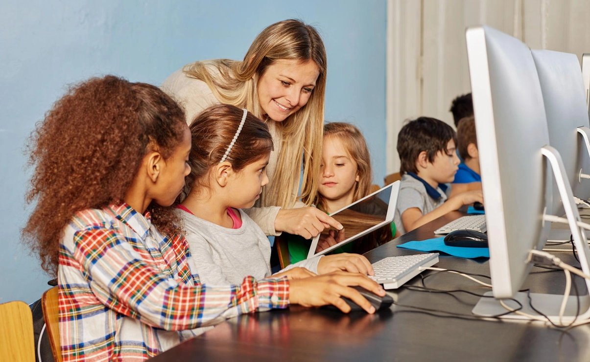 Eine Frau zeigt mehreren Kindern etwas am Tablet. Die Kinder sitzen selbst vor Bildschirmen.