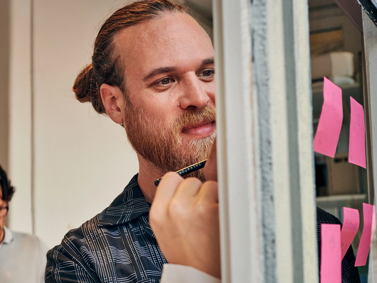 Ein Mann schreibt etwas auf ein Post-It, dass auf einer Durchsichtigen Scheibe klebt.