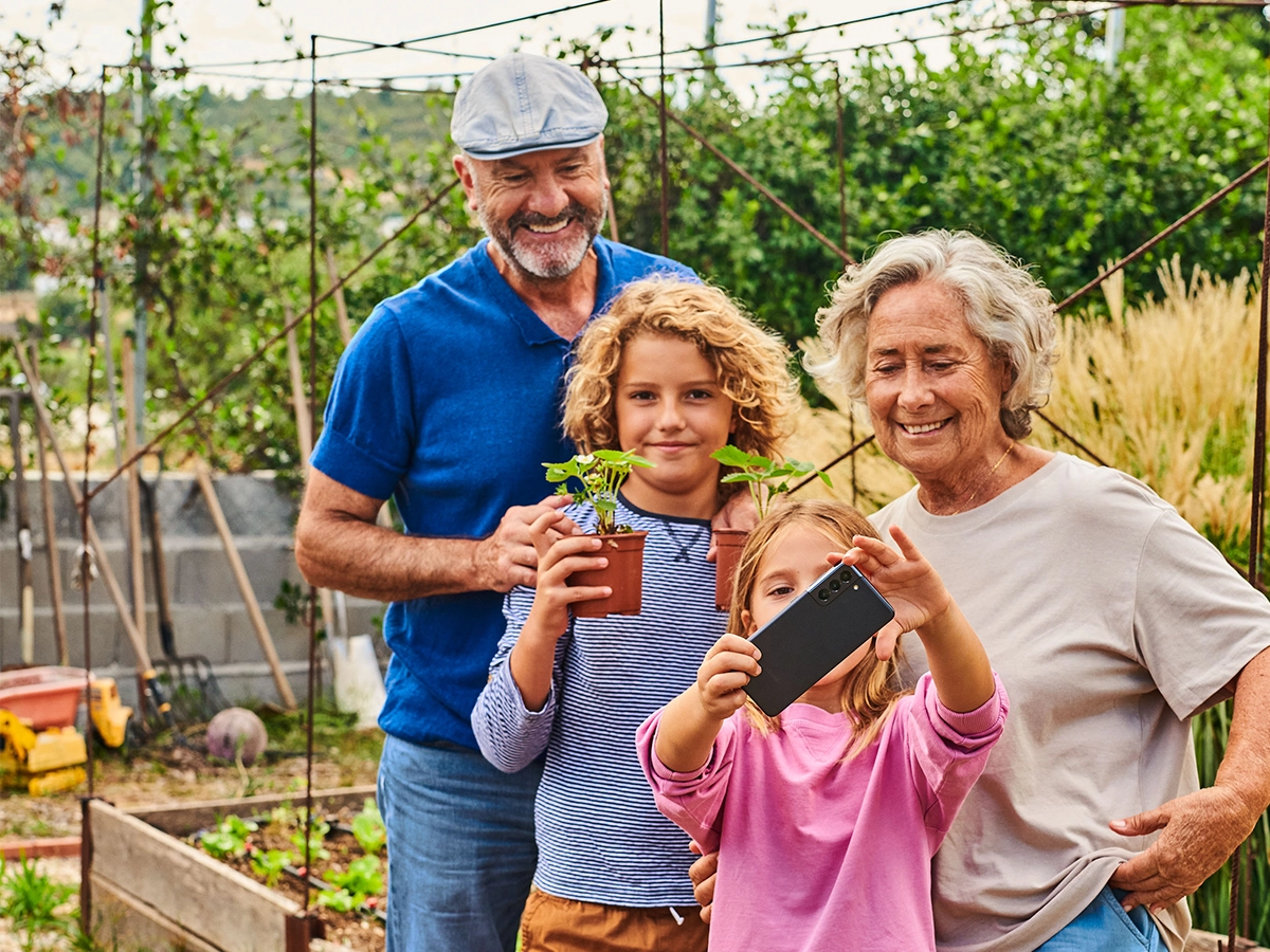 Zwei ältere Menschen und zwei Kinder machen einen Selfie im Garten. Eines der Kinder macht das Selfie während das andere zwei Pflanzen in die Kamera hält.
