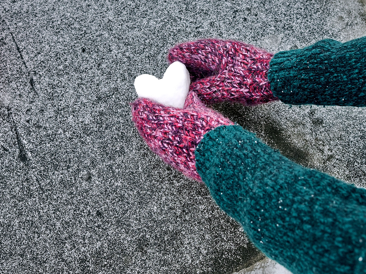 Das Bild zeigt eine Person, die ein aus Schnee geformtes herz auf den Boden legt. Die Person trägt einen Wollpullover und Handschuhe.