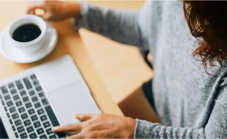 Frau in grauem Pullover trinkt Kaffee beim Arbeiten am Laptop (Foto)