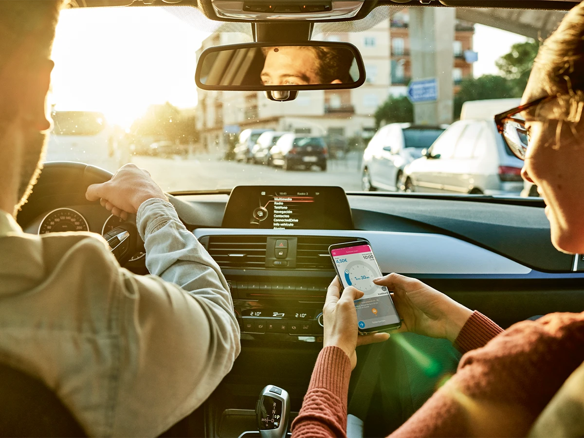 Zwei Personen im Auto, eine Person lenkt, eine blickt auf ein Mobiltelefon. (Header Photo)