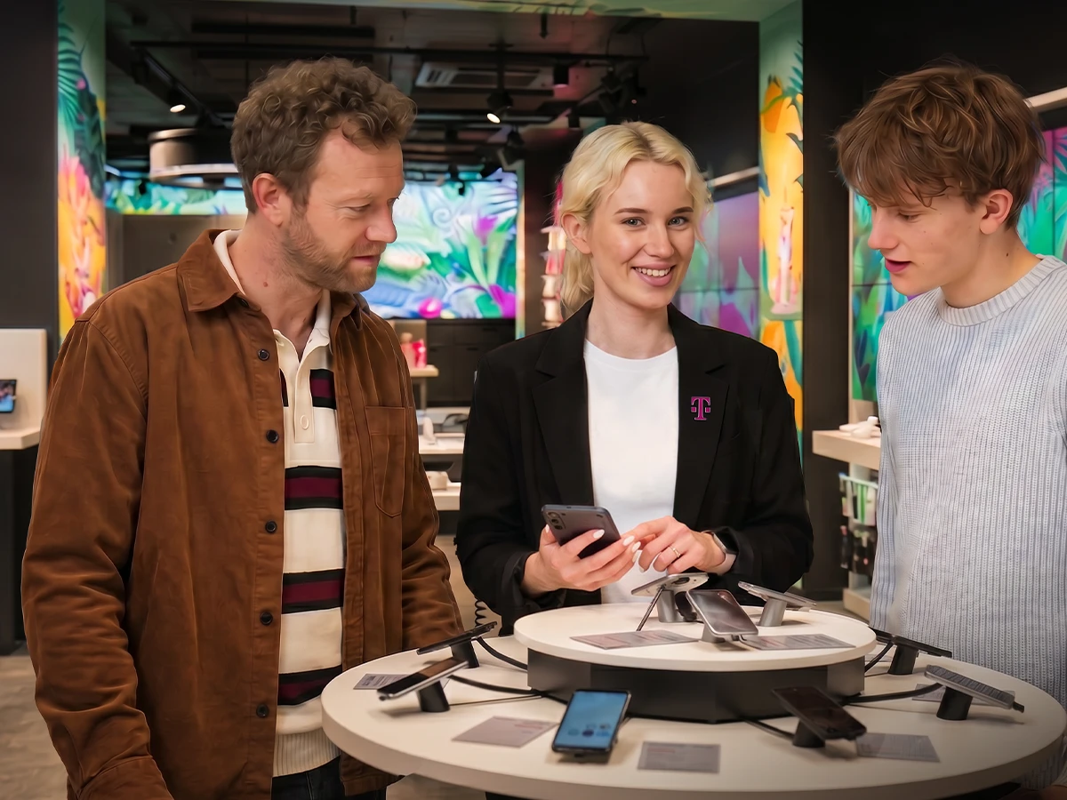 Eine Frau im Telekom Blazer zeigt zwei Männern ein Mobiltelefon in einem Telekom showroom. (Header Photo)