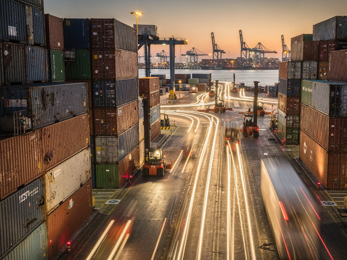 Ein Hafen in der Dämmerung. Links und rechts sind Container gestapelt, im Hintergrund Kräne und in der Mitte eine Straße mit Lichtstreifen von Fahrzeugen (Header Photo)