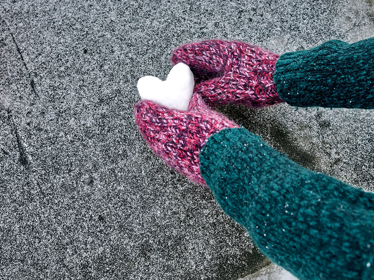 Nahaufnahme einer Person, die ein Herz aus Schnee formt und Fäustline trägt. Man sieht nur Arme und Hände. (Header Photo)