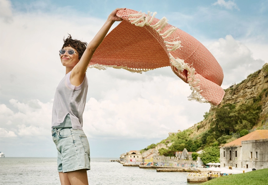 Eine Frau mit Sonnenbrille steht an einer Meeresküste und hält ein orange gemustertes Strandtuch in den Wind. (Foto)