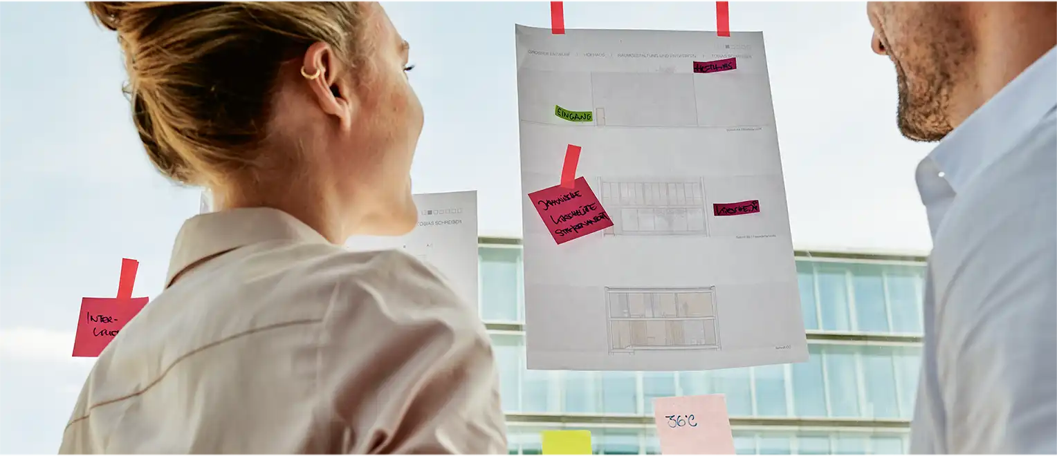 Zwei Personen stehen vor einem Fenster, auf dem verschiedene Post-its kleben (Foto)